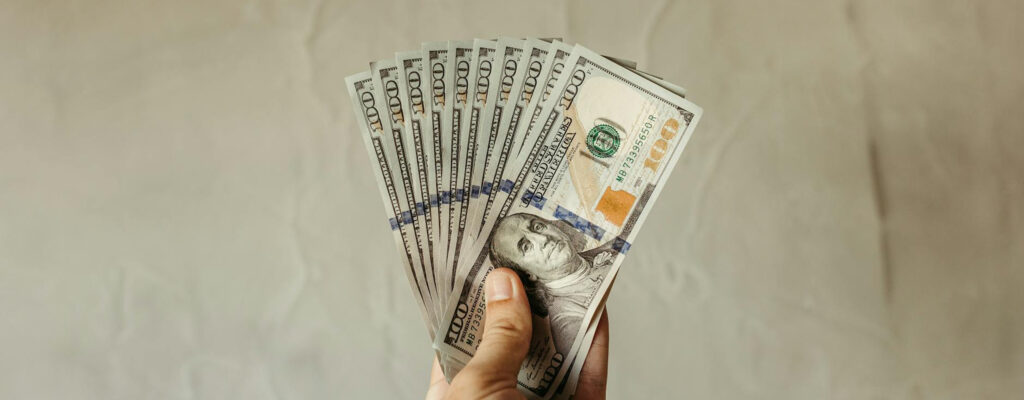 A hand holding a fan of $100 US dollar bills against a beige background.