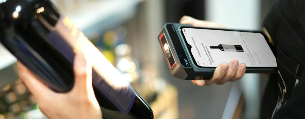 Retail worker scanning a wine bottle using a handheld POS device displaying product information.